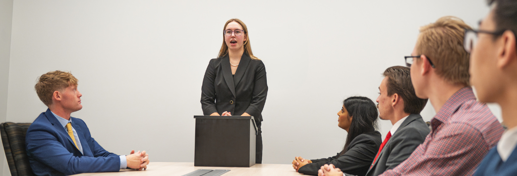 Image of five individuals sitting on either side of a table looking at a young woman speaking behind a podium.